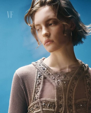 Portrait of a fair-skinned young woman with short brown hair, wearing a beaded top against a soft blue background. Fashion headshot.