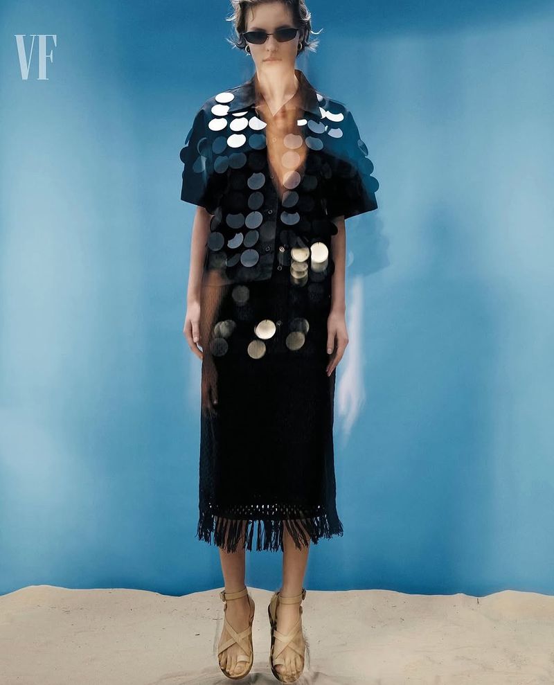 An adult woman stands on a sandy surface in a black outfit with silver circular decorations and strappy sandals against a blue background.