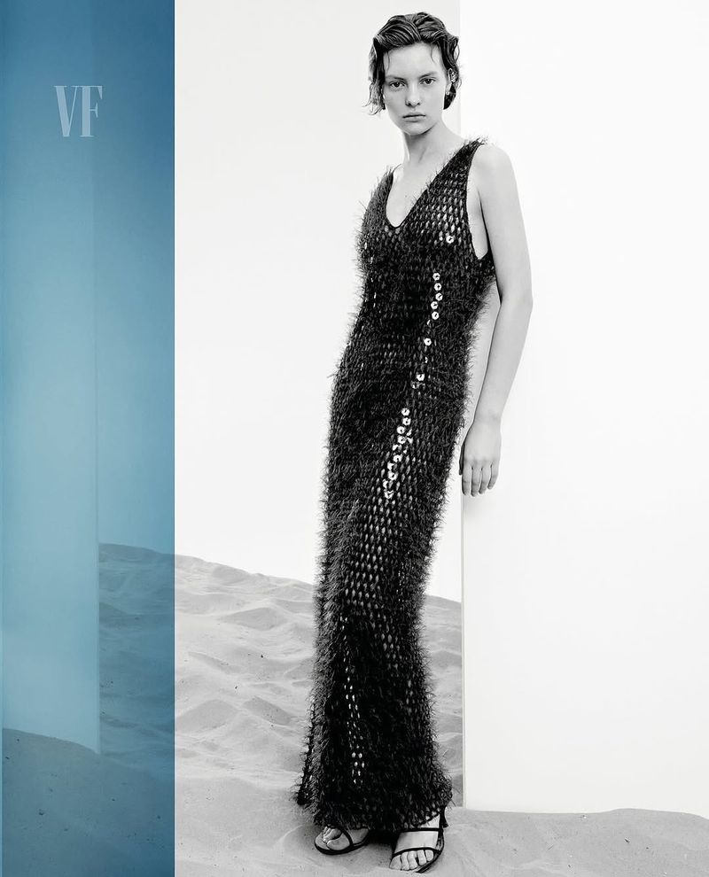 Elegant woman in sequined dress poses near a white wall against a neutral backdrop for a fashion editorial, presented in monochrome.