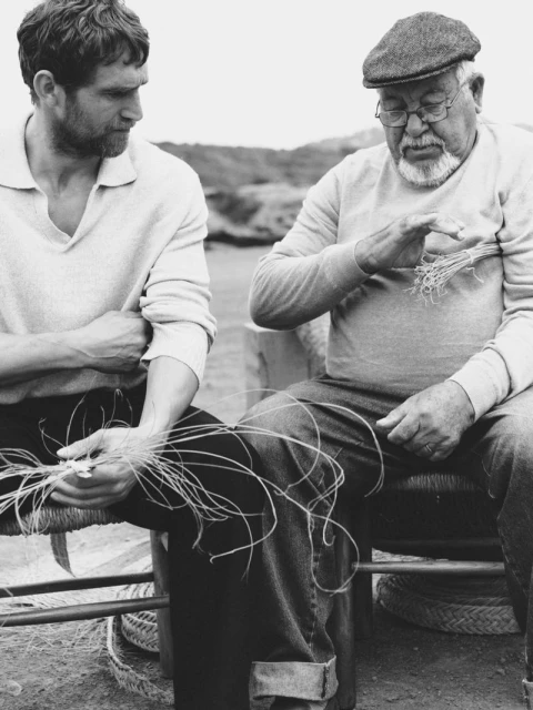 Two men seated outside weaving with natural fibres, wearing casual clothing in a black and white photography style image.