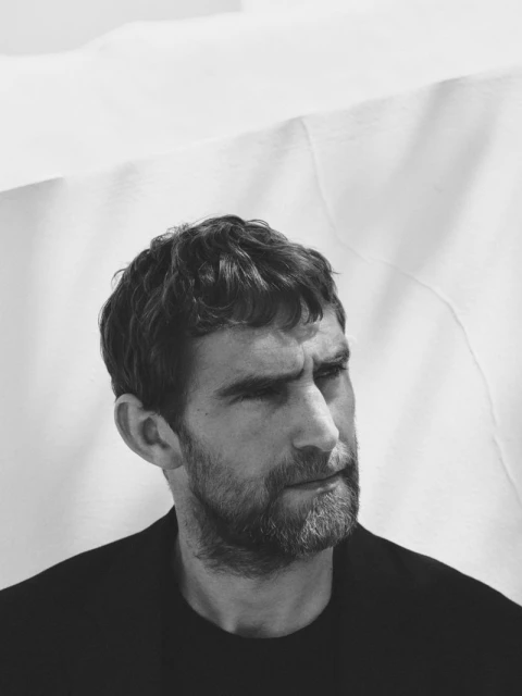 Monochrome portrait of a bearded adult man looking off-camera against a blurred white fabric backdrop, evoking minimalist style.