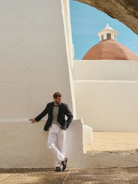 A stylish man in a blazer and white pants leans against a white building, with a terracotta dome visible in the background under a blue sky.