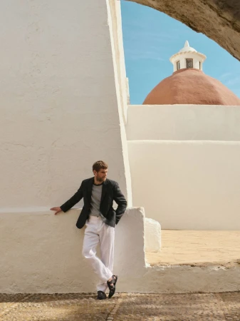 A stylish man in a blazer and white pants leans against a white building, with a terracotta dome visible in the background under a blue sky.