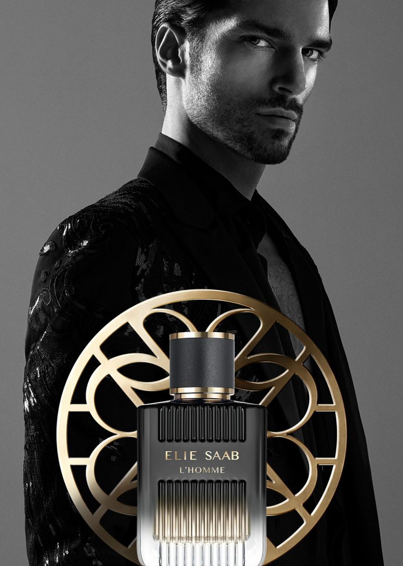 Cologne bottle from Elie Saab, 'L'Homme' in front of a man with a black jacket, ornate gold roundel, and soft grey background. Perfume ad.