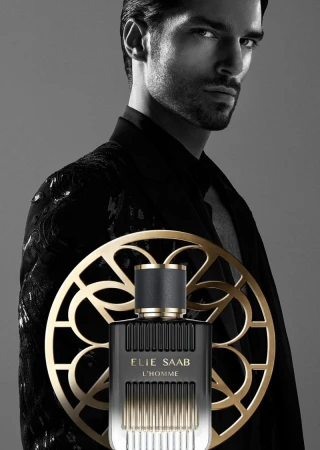 Cologne bottle from Elie Saab, 'L'Homme' in front of a man with a black jacket, ornate gold roundel, and soft grey background. Perfume ad.