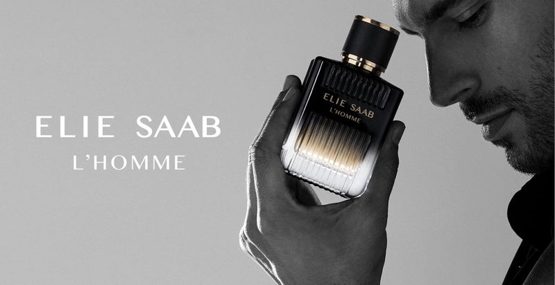 Cologne advertisement featuring a square black and gold bottle of ELIE SAAB L'HOMME held by a man against a gray background.
