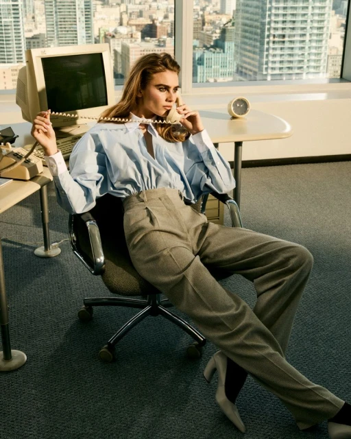 Elegant woman sits in office chair on phone near desktop computer, wearing a blouse and trousers in a modern office setting.