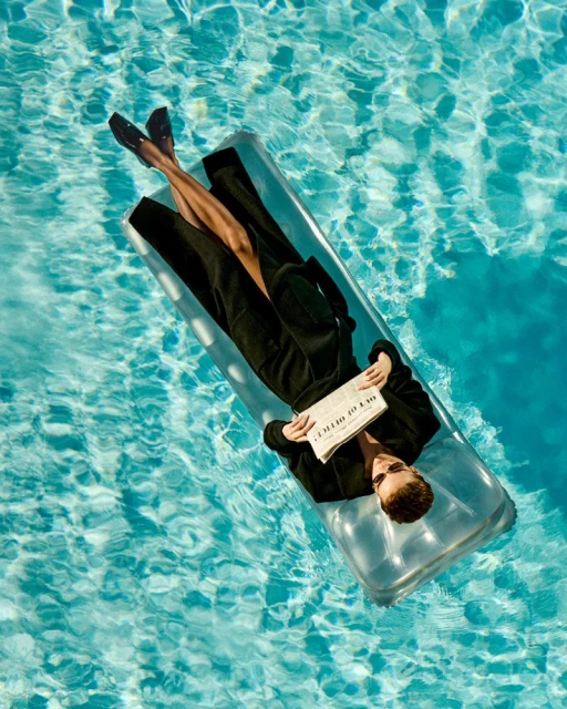 Aerial view of a woman in black on a clear inflatable raft, reading a newspaper in a turquoise swimming pool with dappled sunlight.