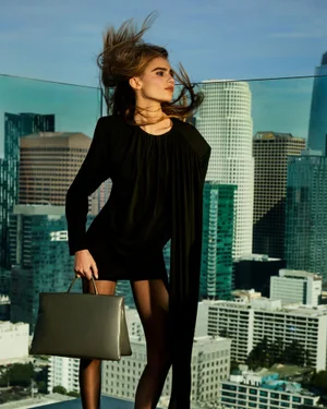 Model in a black dress holding a silver handbag against a city skyline rooftop backdrop