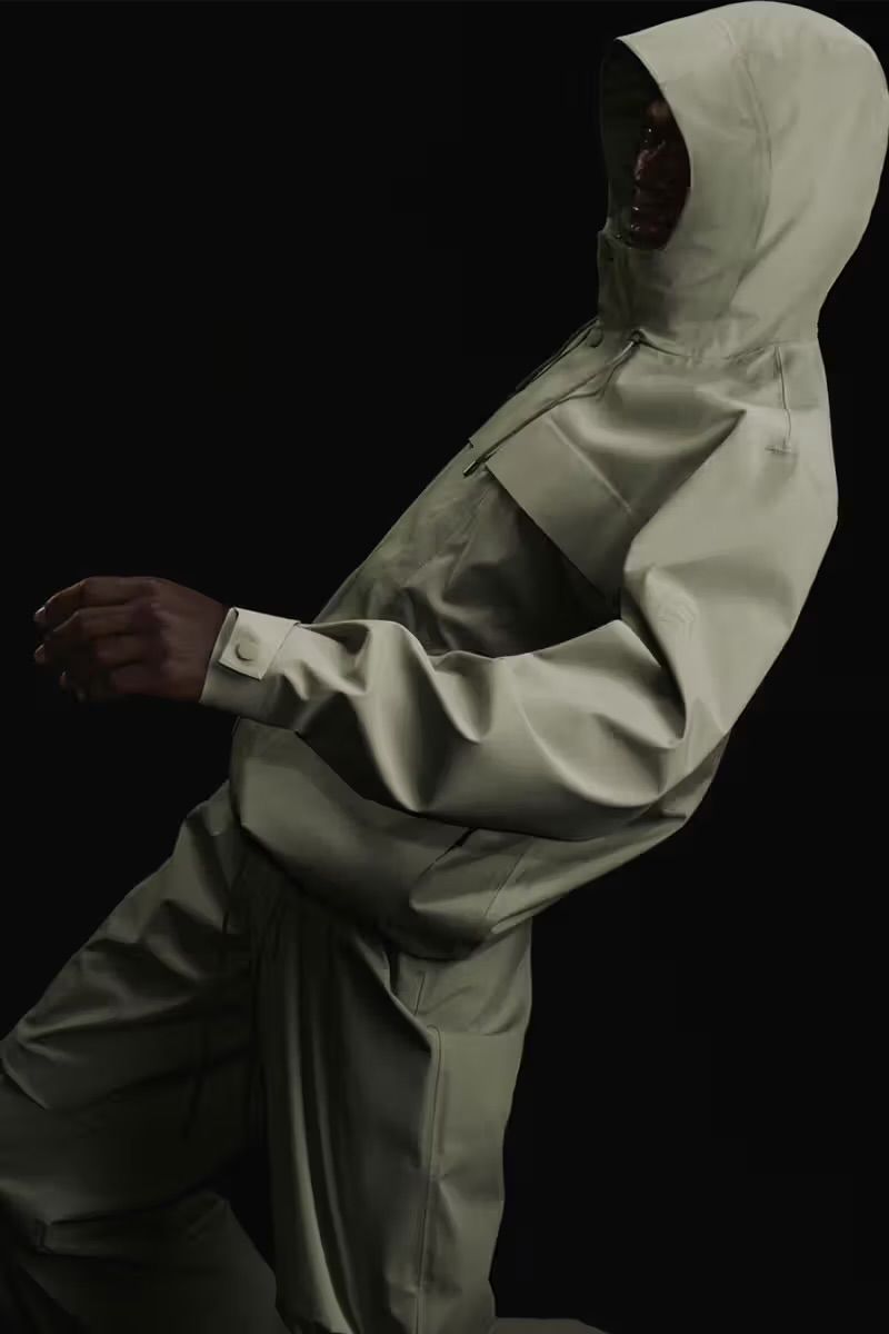 Person wearing a light beige hooded jacket and pants against a dark background in a studio fashion shot.