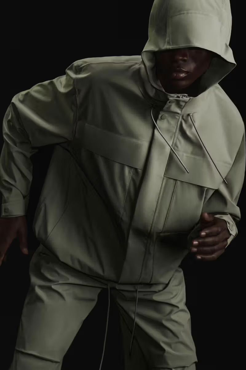 Person in hooded olive-green jacket and pants, against a black background. Minimalist fashion shot. Studio lighting.