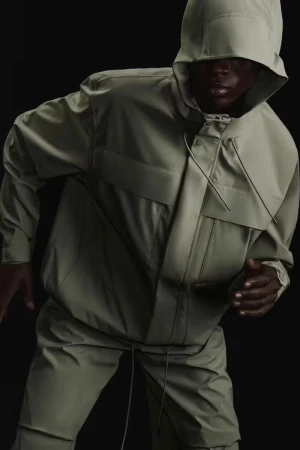 Person in hooded olive-green jacket and pants, against a black background. Minimalist fashion shot. Studio lighting.