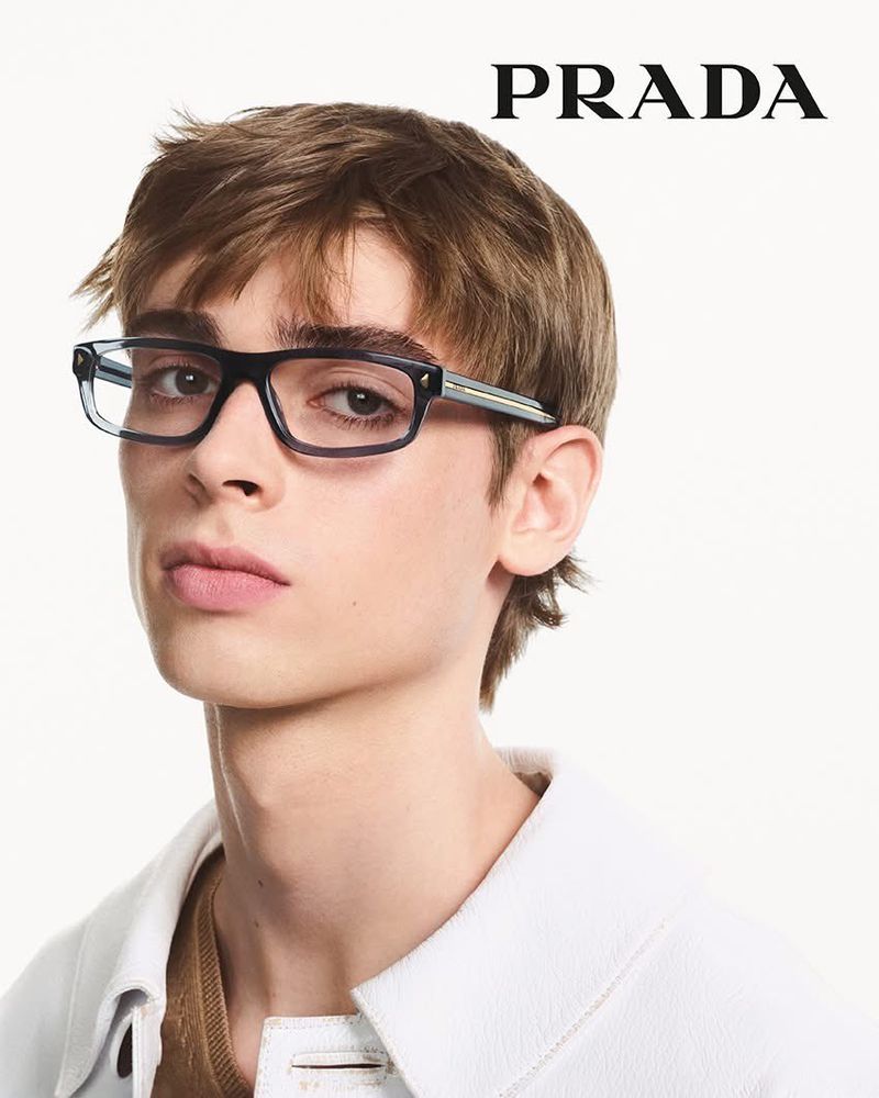 Portrait of a young adult male wearing blue and black framed eyeglasses and a white jacket, with the PRADA logo in the background.