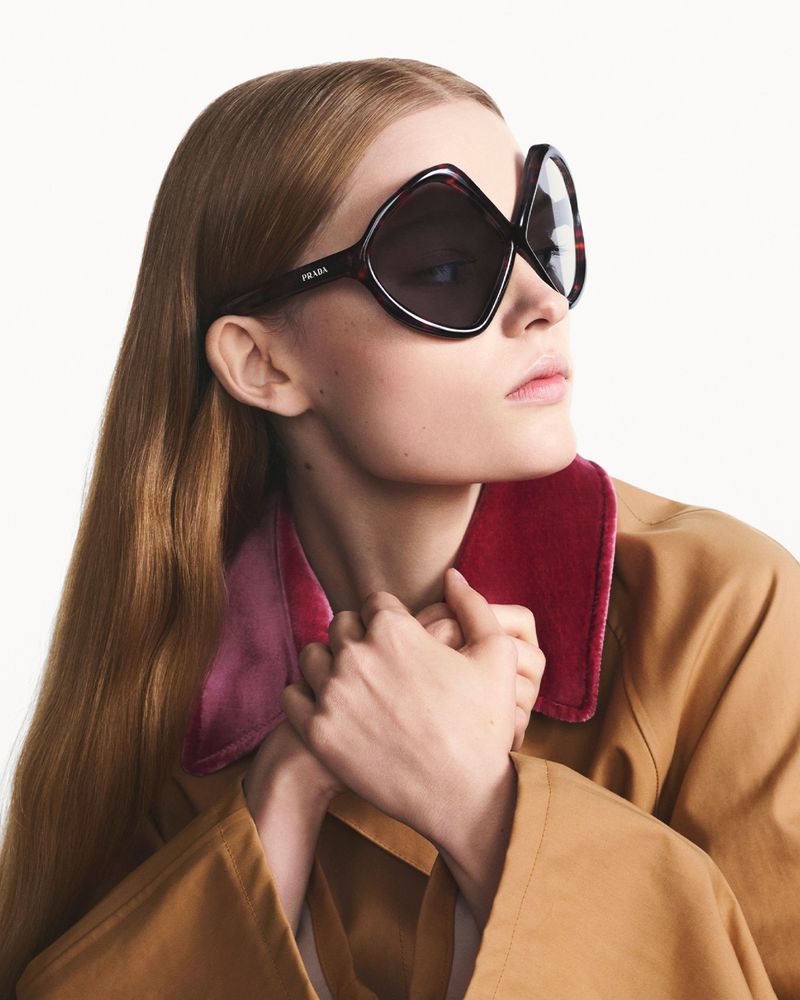 A fashionable adult female with fair skin poses, wearing oversized Prada sunglasses, a tan jacket with a pink collar, against a white backdrop.