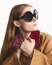 Model wearing oversized tortoiseshell sunglasses and brown jacket against a white background for Prada eyewear campaign