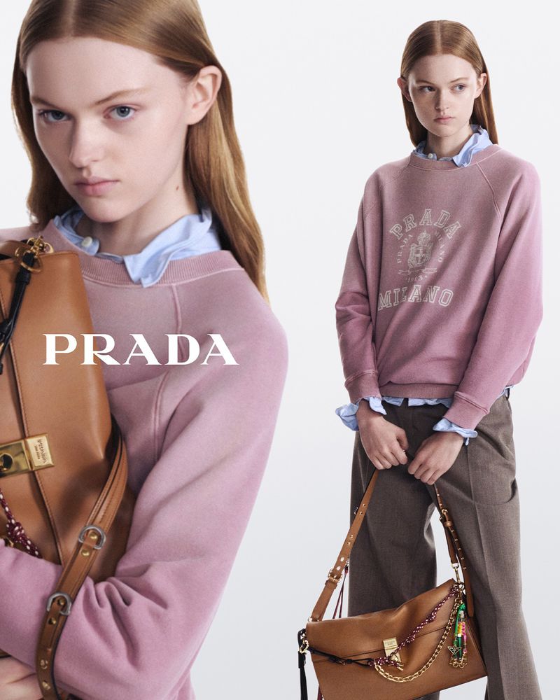 Fashion editorial shot features a redhead woman wearing a pink Prada sweatshirt, paired with brown pants and a brown leather handbag.