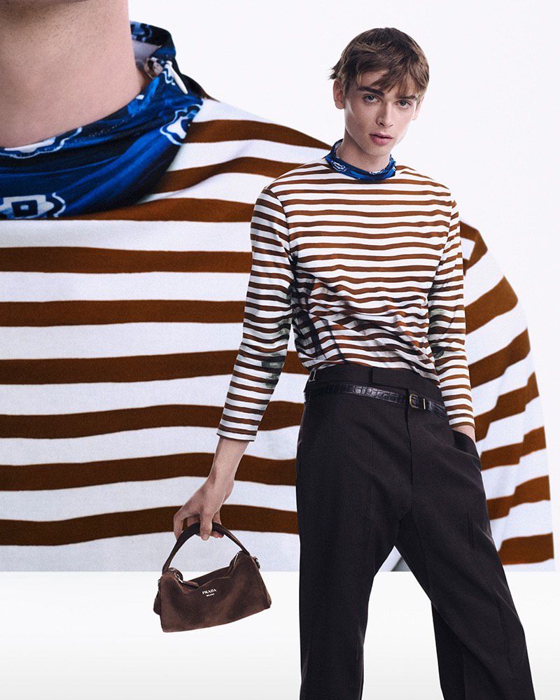 Fashionable adult male in a brown-and-white striped shirt and black pants, holding a brown suede handbag against a white background.