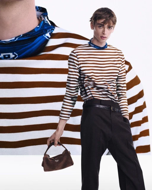 Fashionable adult male in a brown-and-white striped shirt and black pants, holding a brown suede handbag against a white background.