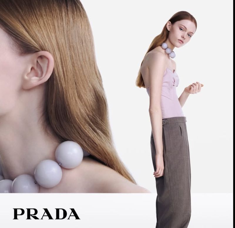 Fashion shot featuring a woman in a lilac top and grey pants wearing a large, round, pale purple beaded necklace against a white backdrop.