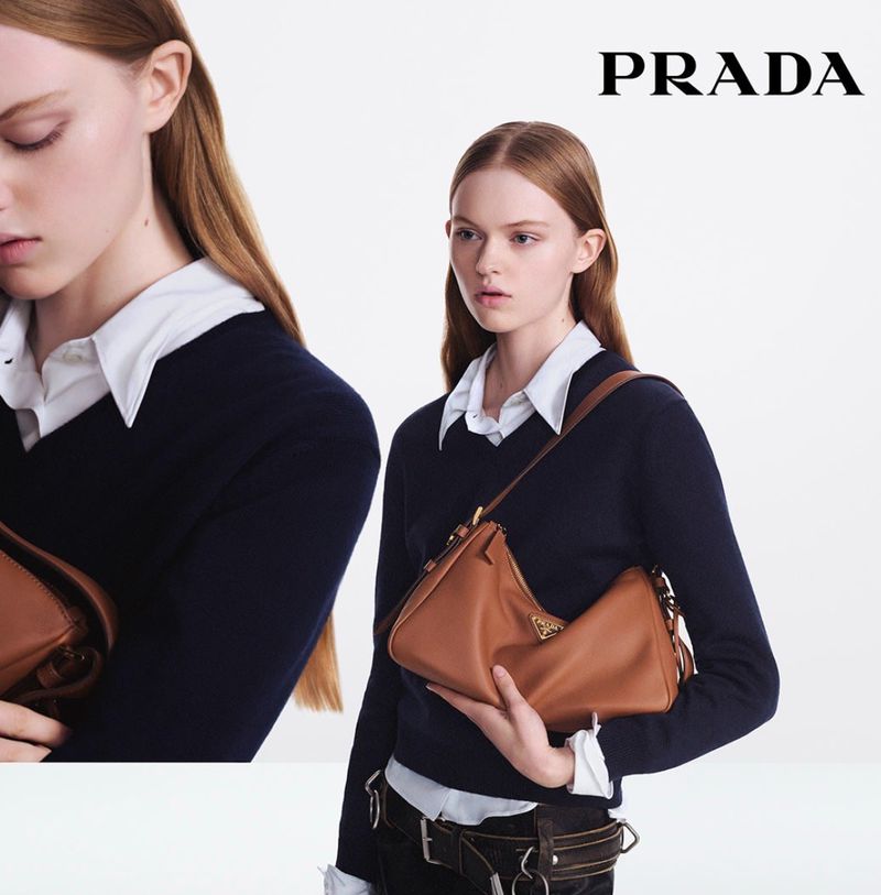 Fashion shot of a woman holding a brown leather Prada handbag, wearing a navy sweater with a white collared shirt on a white background.