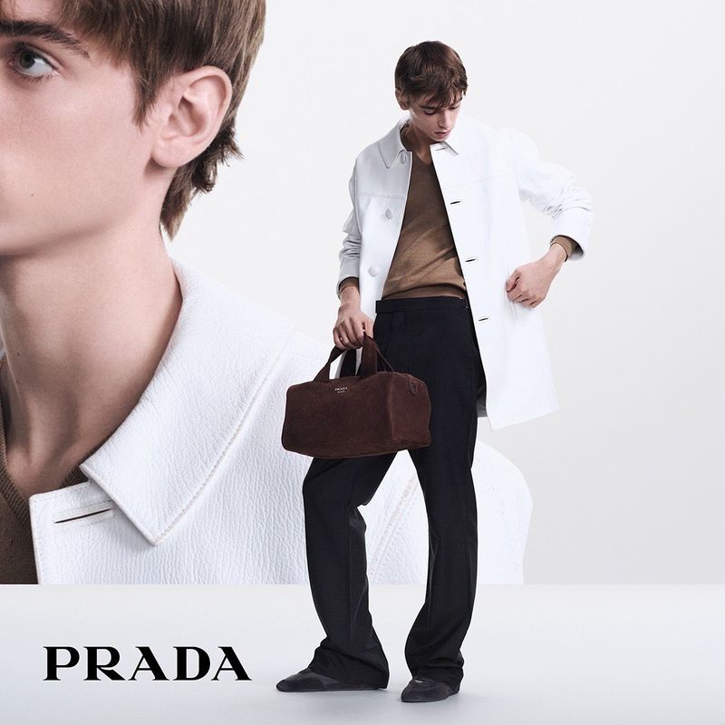 A young adult man in a white coat and black pants is holding a brown leather bag in a fashion-style outdoor studio photo.