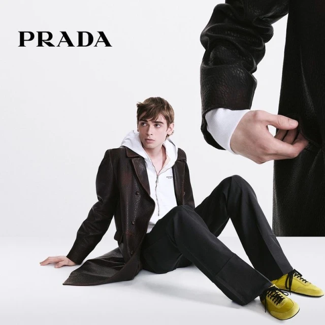 Fashion shot of a young adult male model sitting on a white surface wearing a long brown leather trench coat, white sweatshirt, and yellow sneakers.