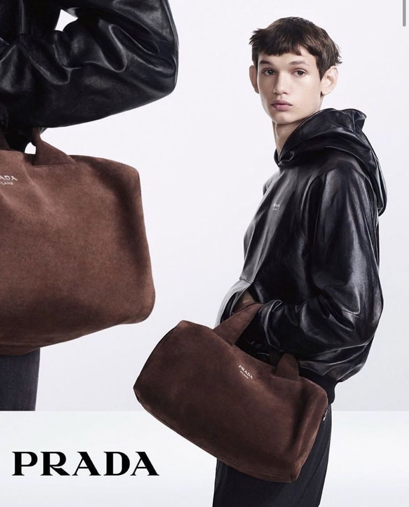 Fashion editorial shot featuring a young male model in a black hooded jacket holding a brown suede Prada tote bag against a white backdrop.