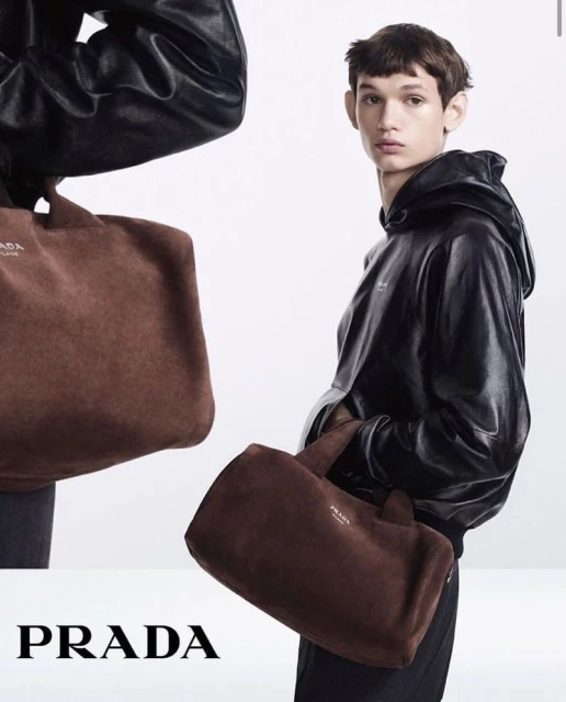 Fashion editorial shot featuring a young male model in a black hooded jacket holding a brown suede Prada tote bag against a white backdrop.