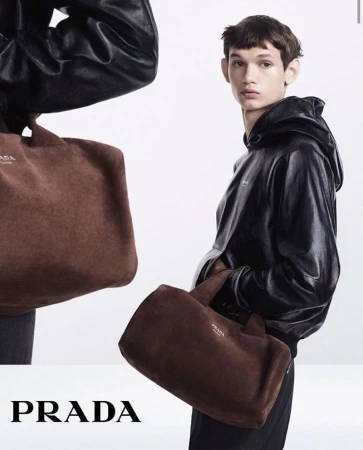 Fashion editorial shot featuring a young male model in a black hooded jacket holding a brown suede Prada tote bag against a white backdrop.