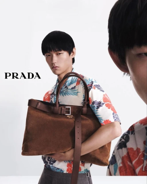 Prada advert featuring an Asian male model holding a brown suede tote bag with leather trim, styled with a floral short-sleeve shirt. 