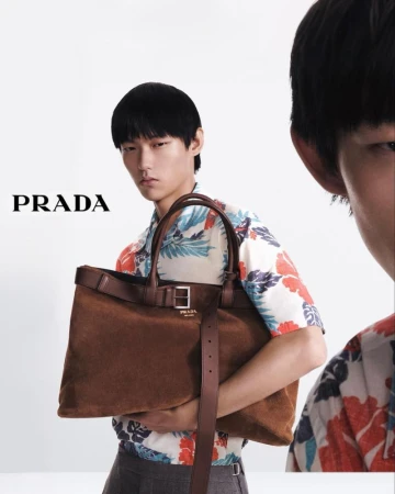 Prada advert featuring an Asian male model holding a brown suede tote bag with leather trim, styled with a floral short-sleeve shirt. 