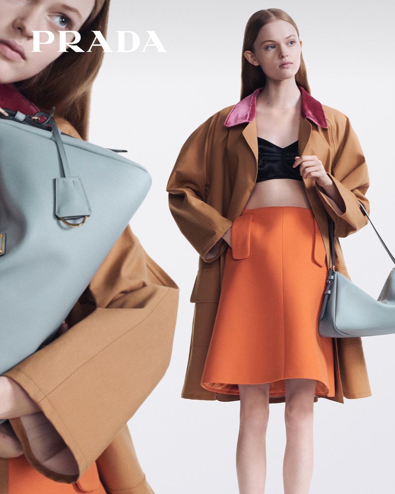 Fashion editorial shot of a woman in skirt and coat holding a Prada handbag against a bright background in a studio setting.