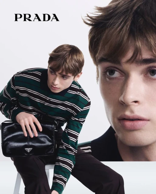 Young man poses with black Prada bag in a studio shot, showcasing a striped top and dark trousers in a fashion-forward context.