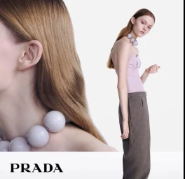 Model wearing a lavender top and grey trousers with a large bead necklace against a white background for Prada S/S 25