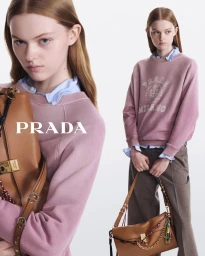 Model wearing a pink crewneck sweatshirt and brown trousers holding a tan leather shoulder bag against a white backdrop