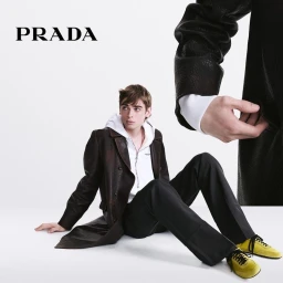 A young male model seated on a white studio floor wearing a brown coat, white hoodie, black trousers, and yellow sneakers.