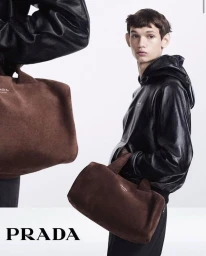 Young man in a black leather hoodie holding a brown suede handbag against a clean white seamless studio background.