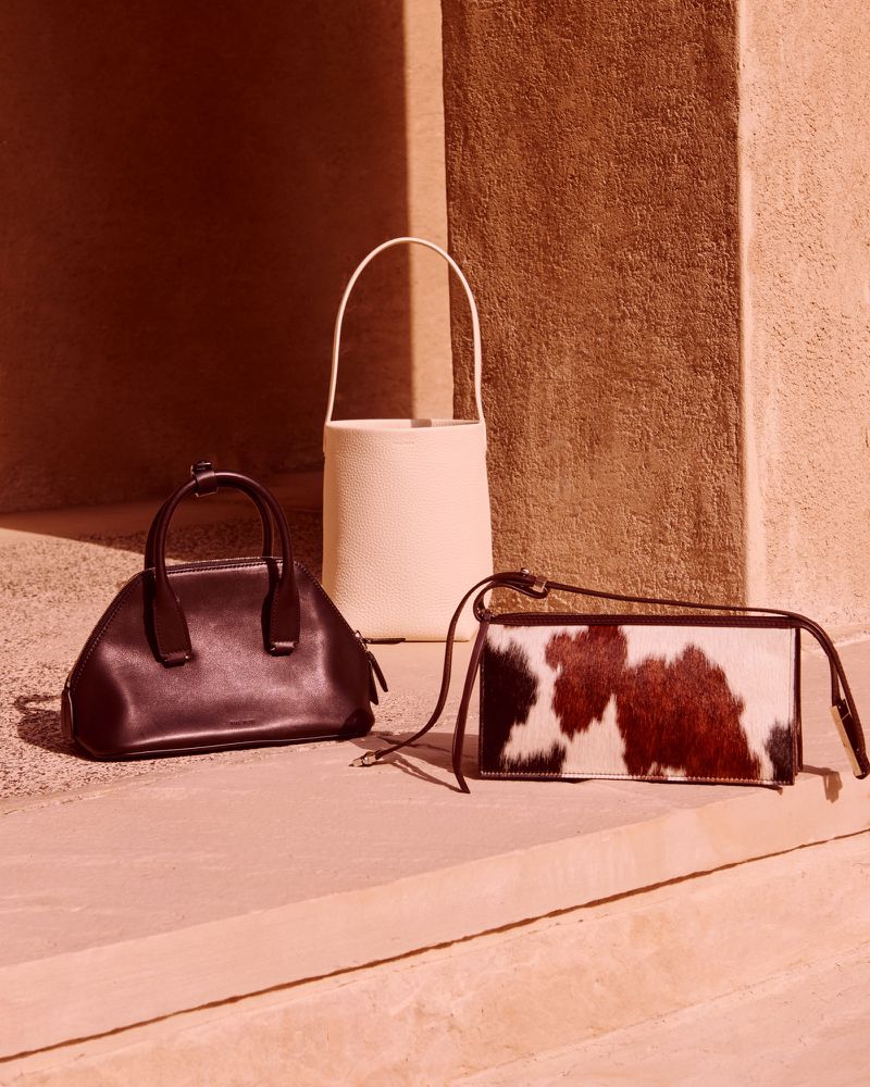 Three designer handbags are displayed indoors on a pink surface with a textured, brown wall providing a simple yet elegant backdrop.