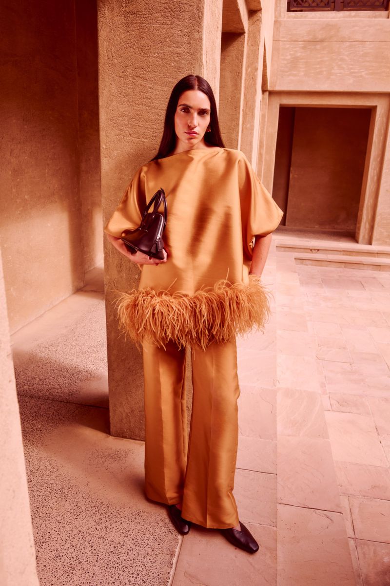 Fashionable woman poses in a peach-toned ensemble featuring a feather-trimmed top and wide-leg pants with a black purse, against an architectural backdrop.