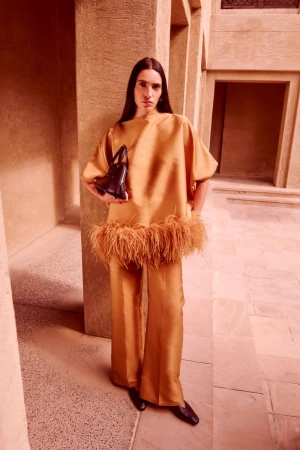 Fashionable woman poses in a peach-toned ensemble featuring a feather-trimmed top and wide-leg pants with a black purse, against an architectural backdrop.