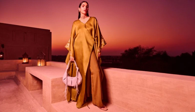 A poised woman models a gold wrap dress with a white purse on a building rooftop with sunset colors in the background.