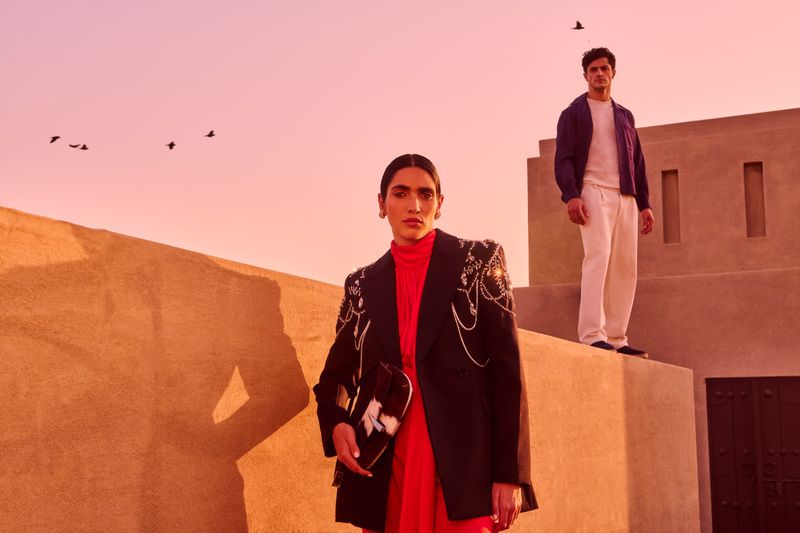 A woman in a black blazer and red dress stands in the foreground, with a man standing on a wall behind her against a pink sunset sky.