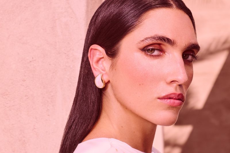 Close-up portrait of a woman with elegant makeup and sleek dark hair, wearing a statement earring, against a soft pink backdrop.