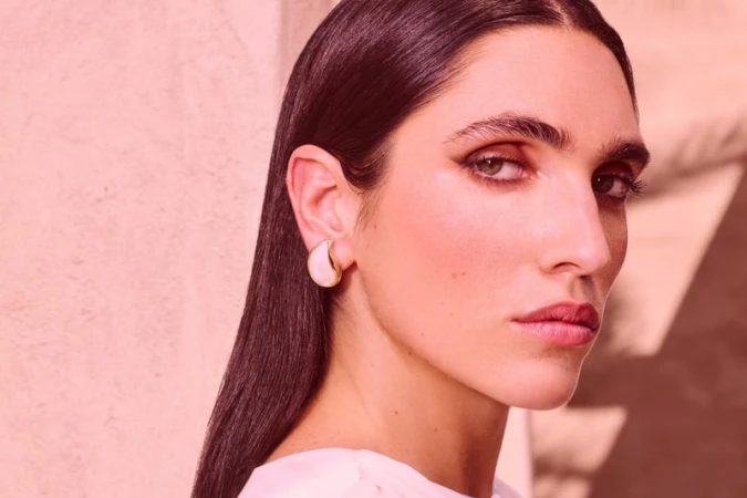 Close-up portrait of a woman with elegant makeup and sleek dark hair, wearing a statement earring, against a soft pink backdrop.