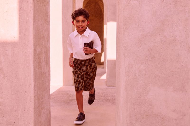 A young boy in designer shorts and a white shirt smiles while running through a pink-toned corridor with open doorways and bright sunlight.