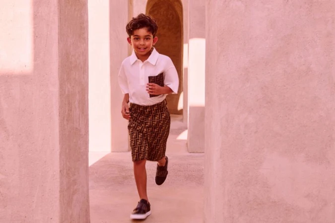 A young boy in designer shorts and a white shirt smiles while running through a pink-toned corridor with open doorways and bright sunlight.