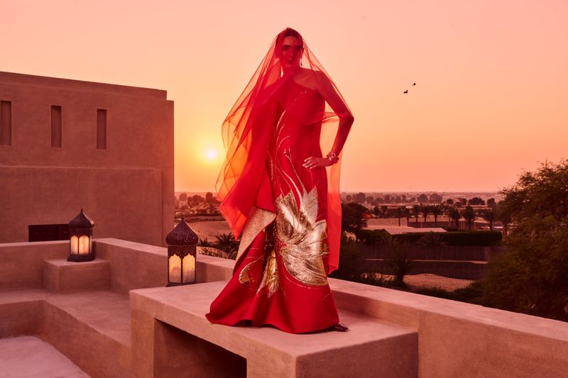 A woman in a red gown and veil poses on a rooftop at sunset, with warm lighting and a desert landscape in the background.