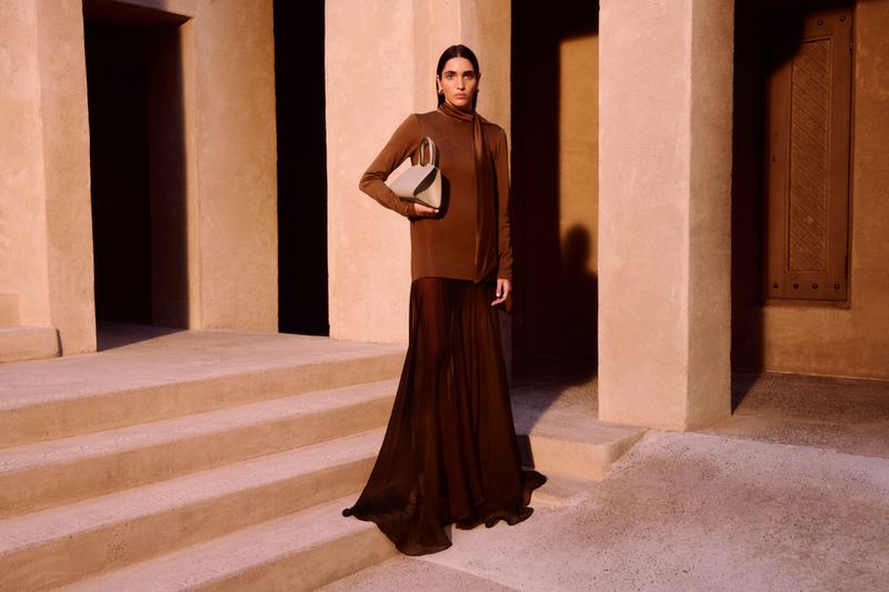 Fashion shoot of a model in a long-sleeved brown dress and a tan and green handbag, posing on outdoor steps, warm color palette.