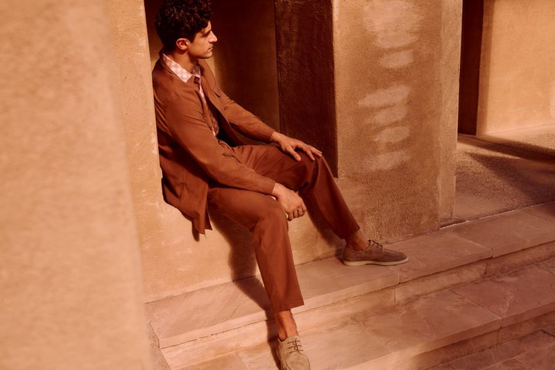 Adult male model poses while sitting on a sandstone step, wearing a brown suit and casual shoes. The light is soft, and the background is architectural.
