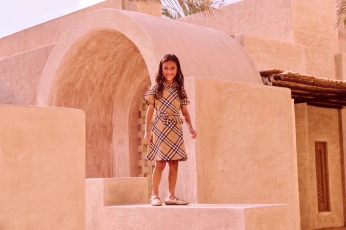 Young girl in plaid dress smiles on architectural steps in warm sunlight. Fashion shot with desert environment in the background.
